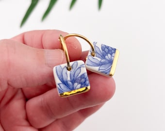 May include: A pair of gold hoop earrings with blue and white porcelain square charms. The charms feature a floral design with gold trim.