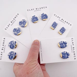 Könnte beinhalten: Vier Paar blaue und weiße Blumen-Ohrringe aus Keramik mit goldenen Akzenten. Die Ohrringe werden auf weißen Karten mit dem Text "Clay Blanca Ceramics" und "Handmade in Yorkshire" präsentiert.