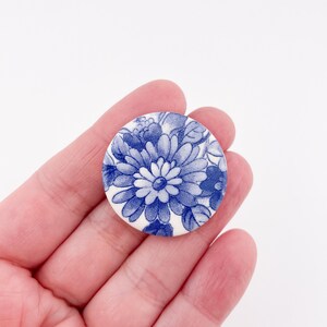 May include: A round, white button with a blue floral design. The button features a detailed pattern of flowers and leaves in shades of blue, creating a classic, vintage aesthetic. The button is held in a person's hand.