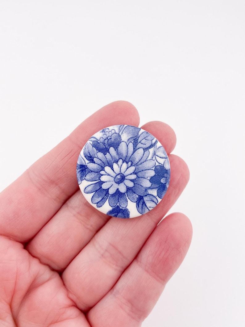 May include: A round, white button with a blue floral design. The button features a detailed pattern of flowers and leaves in shades of blue, creating a classic, vintage aesthetic. The button is held in a person's hand.
