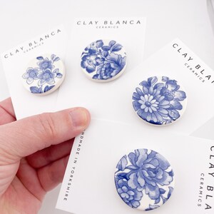May include: Four round ceramic brooches with blue floral designs on white backgrounds. Each brooch is mounted on a white card with the text "CLAY BLANCA CERAMICS" and "HANDMADE IN YORKSHIRE". The brooches feature different blue floral patterns.