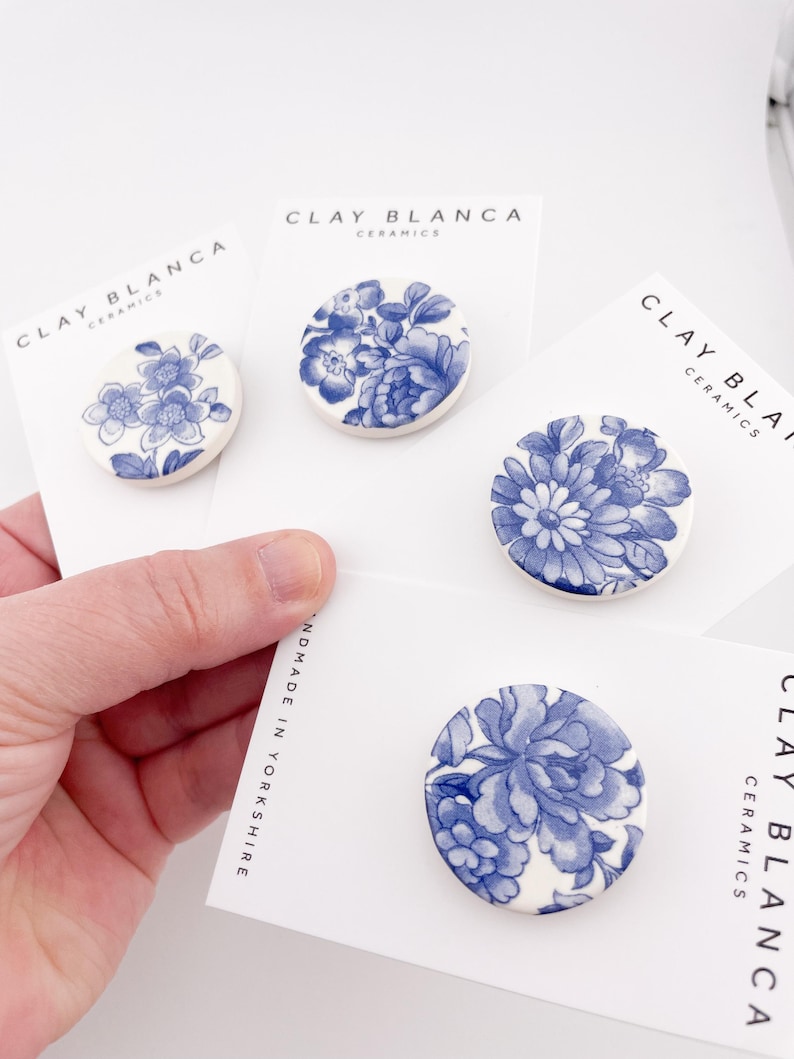 May include: Four round ceramic brooches with blue floral designs on white backgrounds. Each brooch is mounted on a white card with the text "CLAY BLANCA CERAMICS" and "HANDMADE IN YORKSHIRE". The brooches feature different blue floral patterns.