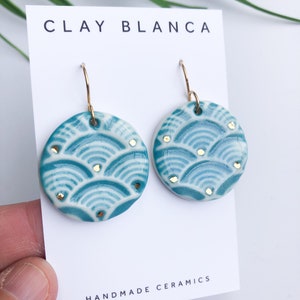 Op de afbeelding: Turquoise blauwe keramische oorbellen met een gouden geometrisch patroon en gouden haakjes. De oorbellen worden getoond op een witte kaart met de tekst "CLAY BLANCA HANDMADE CERAMICS".