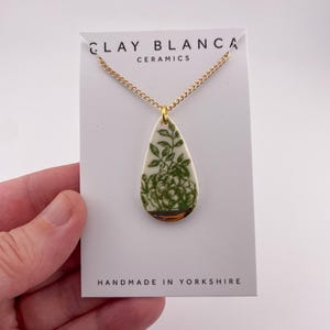 Op de afbeelding: Een ketting met een keramische hanger in de vorm van een druppel. De hanger heeft een groen bloemmotief op een witte achtergrond, met een gouden rand. De kaart leest "CLAY BLANCA CERAMICS" en "HANDMADE IN YORKSHIRE."