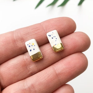 Puede incluir: Un par de pendientes de botón de cerámica blanca con detalles dorados y motas azules y amarillas.