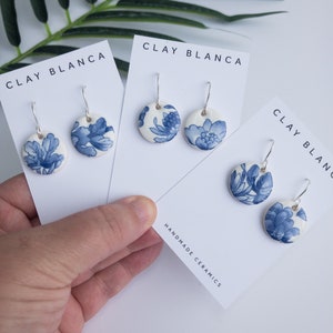 Könnte beinhalten: Drei Paar blaue und weiße Blumen-Keramikohrringe mit silbernen Haken. Die Ohrringe werden auf weißen Karten mit dem Text "CLAY BLANCA" und "HANDMADE CERAMICS" präsentiert.