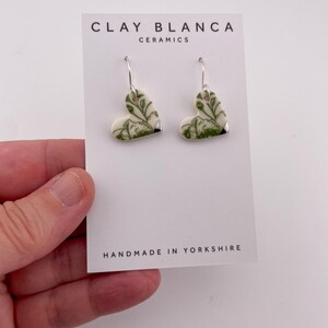 Op de afbeelding: Hartvormige keramische oorbellen met een groen botanisch ontwerp en zilveren haken. De oorbellen worden gepresenteerd op een witte kaart met de tekst "CLAY BLANCA CERAMICS" en "HANDMADE IN YORKSHIRE".
