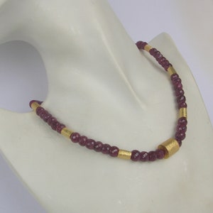 Puede incluir: Un collar hecho de pequeñas cuentas de color rojo oscuro con detalles dorados. El collar está en un maniquí blanco.