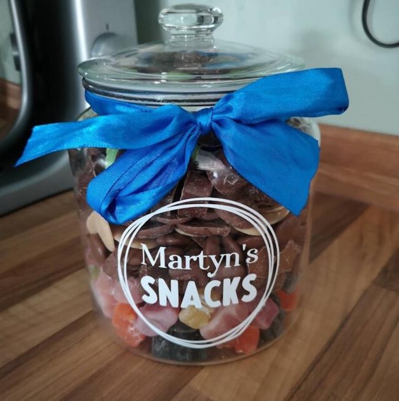 May include: A clear glass jar with a lid, filled with assorted brown chocolates and colorful candies. A bright blue ribbon is tied around the neck of the jar. The front features a white circular graphic with the text Martyn's SNACKS.