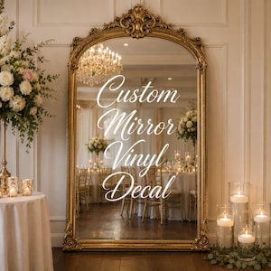 May include: A large, arched floor mirror with an ornate gold frame. The glass surface features white vinyl lettering that reads Custom Mirror Vinyl Decal in a cursive script font. The mirror reflects a formal event space with tables and chairs.