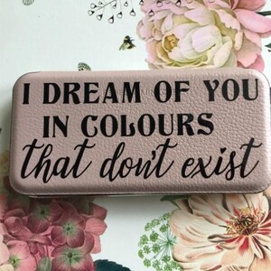 May include: A rectangular, dusty pink wallet with a textured surface. The front features black text that reads: I DREAM OF YOU IN COLOURS that don't exist. The wallet has rounded corners and a slim, compact design with a visible zipper along the edge.
