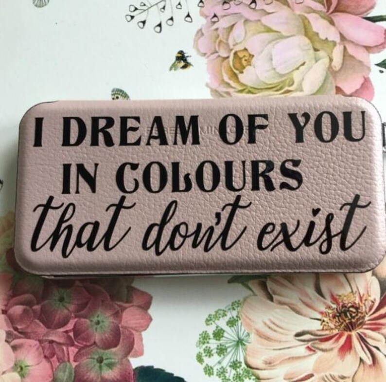 May include: A rectangular, dusty pink wallet with a textured surface. The front features black text that reads: I DREAM OF YOU IN COLOURS that don't exist. The wallet has rounded corners and a slim, compact design with a visible zipper along the edge.