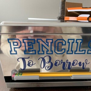 May include: A rectangular, reflective silver metal storage box featuring the blue text PENCILS To Borrow on the front. Two yellow pencils with pink erasers are resting horizontally in a gray plastic holder attached to the front of the box.
