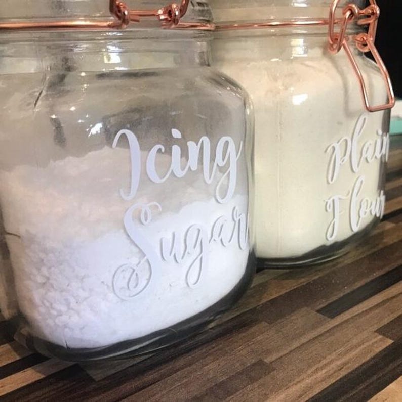 May include: Two clear glass storage jars with copper-colored metal wire-bail lids. The front jar features white cursive text reading Icing Sugar, while the back jar displays white cursive text reading plain flour. Both jars are filled with white powder.