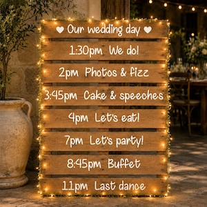 May include: A wooden pallet wedding day schedule sign featuring white text that reads: Our wedding day, 1:30pm We do!, 2pm Photos & fizz, 3:45pm Cake & speeches, 4pm Let's eat!, 7pm Let's party!, 8:45pm Buffet, 11pm Last dance, framed by warm fairy lights.