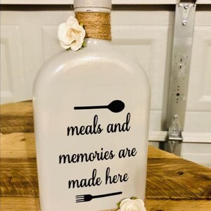May include: A white decorative bottle featuring a black graphic of a spoon, the text "meals and memories are made here," and a black graphic of a fork. The neck of the bottle is wrapped in brown twine and adorned with two white fabric flowers.