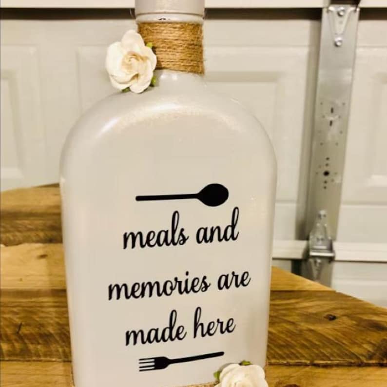May include: A white decorative bottle featuring a black graphic of a spoon, the text "meals and memories are made here," and a black graphic of a fork. The neck of the bottle is wrapped in brown twine and adorned with two white fabric flowers.
