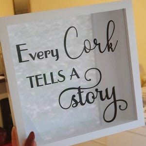 May include: A square white shadow box frame with a glass front. The glass features black lettering that reads Every Cork TELLS A story in a mix of serif and script fonts. The frame is empty, showing a clear view through the glass pane.