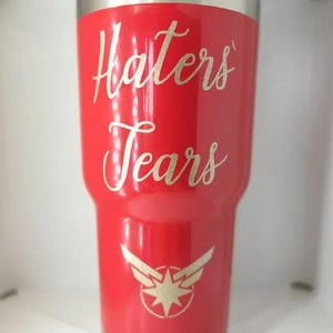 May include: A glossy red stainless steel tumbler featuring the gold glitter text Haters Tears in a cursive script font. Below the text is a gold glitter graphic of a stylized eight-pointed star emblem with wings extending from the sides.