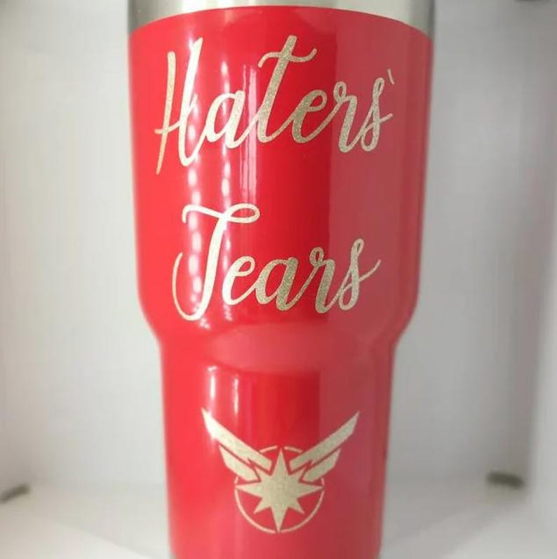 May include: A glossy red stainless steel tumbler featuring the gold glitter text Haters Tears in a cursive script font. Below the text is a gold glitter graphic of a stylized eight-pointed star emblem with wings extending from the sides.