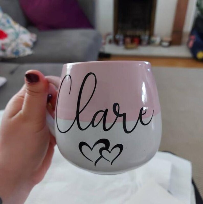 May include: A rounded ceramic mug featuring a two-tone design with a soft pink upper section and a white lower section. The name Clare is printed in black cursive script across the center, accompanied by two black heart outlines below the text.