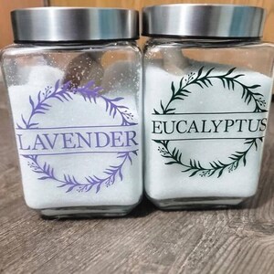 May include: Two square glass jars with silver metal lids, filled with white granular contents. The left jar features a purple laurel wreath graphic with the text LAVENDER. The right jar features a dark green laurel wreath graphic with the text EUCALYPTUS.