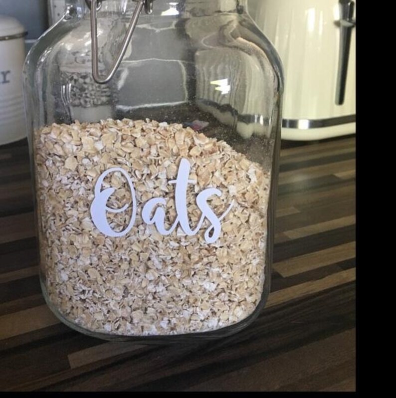 May include: A clear glass storage jar with a metal wire bail closure, filled with dry rolled oats. The front of the jar features the word Oats written in a white, cursive script font. The container is rectangular with rounded corners.