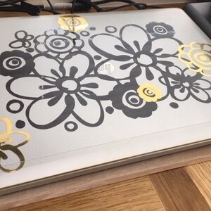 May include: A silver laptop lid decorated with a large floral decal featuring black and metallic gold flower outlines and circular patterns. The design covers the center and corners of the surface, showcasing a mix of solid black and shiny gold petals.