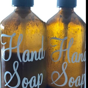 May include: Two amber glass bottles with black screw-top caps, each featuring the white cursive text Hand Soap printed on the front. The bottles are filled with a dark liquid, and the internal pump tubes are visible through the glass.