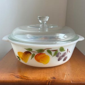 May include: A white, round casserole dish with a clear glass lid. The dish is decorated with hand-painted yellow and orange peaches, green leaves, and purple flowers. The dish has two handles and a clear glass knob on the lid.