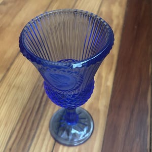May include: A cobalt blue glass goblet featuring a flared rim with vertical ribbing, a central band with a diamond-cut pattern, and a circular medallion depicting a bird in flight. The stem is short and connects to a round, rayed base.
