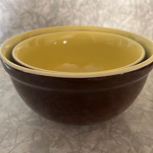 May include: Two nested mixing bowls. The larger bowl is a deep brown with a cream-coloured rim. The interior of the larger bowl is light yellow. The smaller bowl is light yellow with a cream-coloured rim and fits inside the larger bowl.