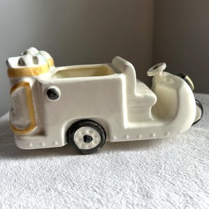 May include: A white ceramic planter shaped like a vintage car. The planter has black wheels, a steering wheel, and a yellow accent around the top and sides. The car-shaped planter is designed to hold small plants or flowers.