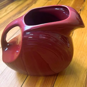 May include: A red ceramic pitcher with a wide mouth and a curved handle. The pitcher has a glossy finish and a smooth, rounded shape.