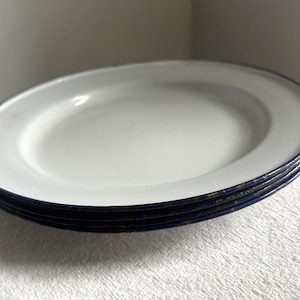 May include: A stack of four white enamel plates with a dark blue rim. The plates are oval-shaped and have a slightly distressed appearance, suggesting a vintage or rustic aesthetic. The plates are stacked on a white textured surface.