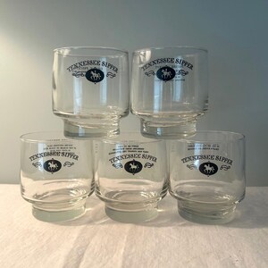 May include: A stack of five clear glass tumblers. Each tumbler has a wide base and a cylindrical body. The text "Tennessee Sipper" is printed on each glass, along with a horse and rider emblem.