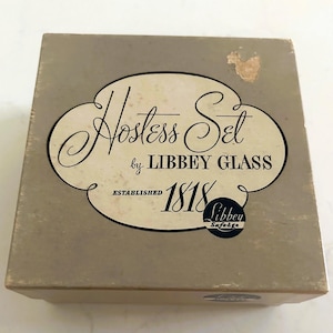 May include: A vintage Hostess Set box by Libbey Glass, established in 1818. The box is a light tan color with a white and black oval label that reads "Hostess Set". The Libbey logo is also visible.