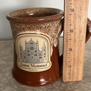 May include: A brown ceramic mug with a white drip glaze at the rim, featuring a black line drawing of the Saint Meinrad Archabbey in Indiana. A wooden ruler placed next to the mug shows it stands approximately 4 inches tall.