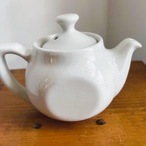 May include: A small, white ceramic teapot with a matching lid. The teapot has a rounded body, a curved handle, and a spout. The lid has a small knob for easy removal. The teapot is sitting on a wooden surface.