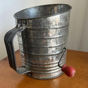 May include: Vintage metal flour sifter with a black handle and a red wooden crank. The cylindrical sifter has a distressed, aged appearance, with horizontal ridges around the body. A classic kitchen tool.