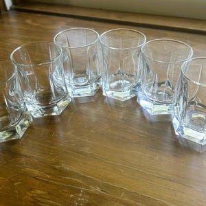 May include: A set of six clear glass tumblers, each featuring a distinct hexagonal base that transitions into a rounded rim. The glasses are made of transparent, smooth glass with vertical facets along the lower portion of the body, creating a geometric design.