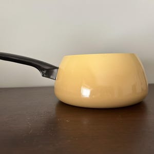 May include: A round, light yellow saucepan with a smooth, glossy finish and a single, long, black handle extending from the side. The pot features a wide, curved body and a flat base, designed for stovetop cooking with a simple, functional aesthetic.