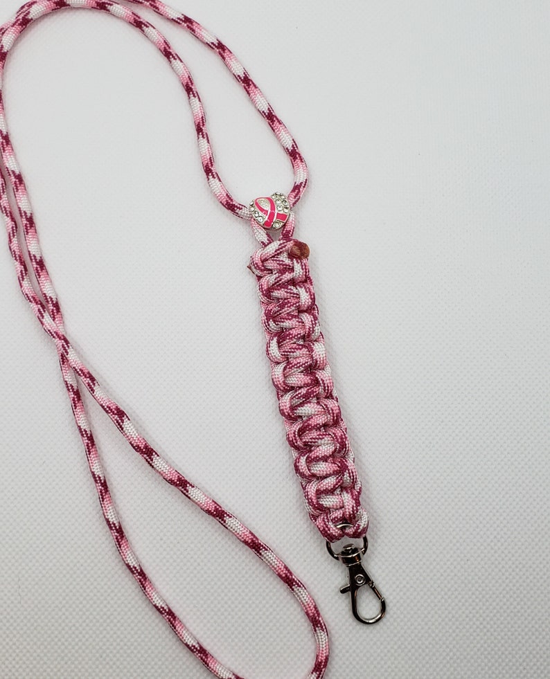 May include: A pink and white lanyard with a braided section and a silver-colored clasp. The lanyard features a pink ribbon charm with rhinestone accents, symbolizing breast cancer awareness. The cord is a mix of pink, white, and burgundy.
