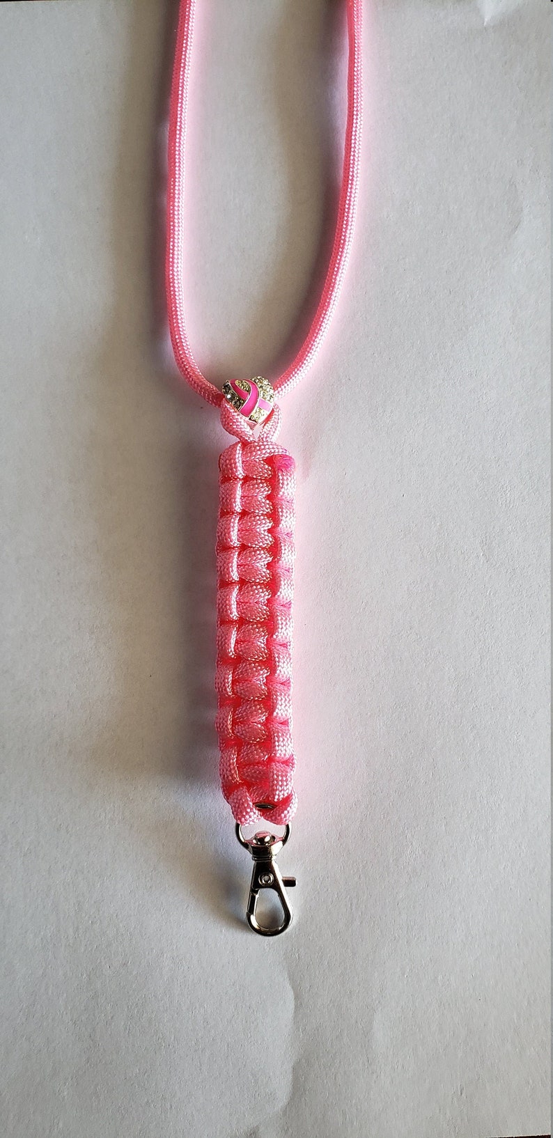 May include: Pink lanyard with a woven design, featuring a silver-colored clasp. The lanyard has a pink ribbon with a heart-shaped charm with a pink ribbon design. The lanyard is approximately 18 inches long.
