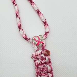 May include: A pink and white braided lanyard with a pink ribbon charm. The charm is encrusted with rhinestones and has a pink ribbon in the center. The lanyard is made of a braided cord.