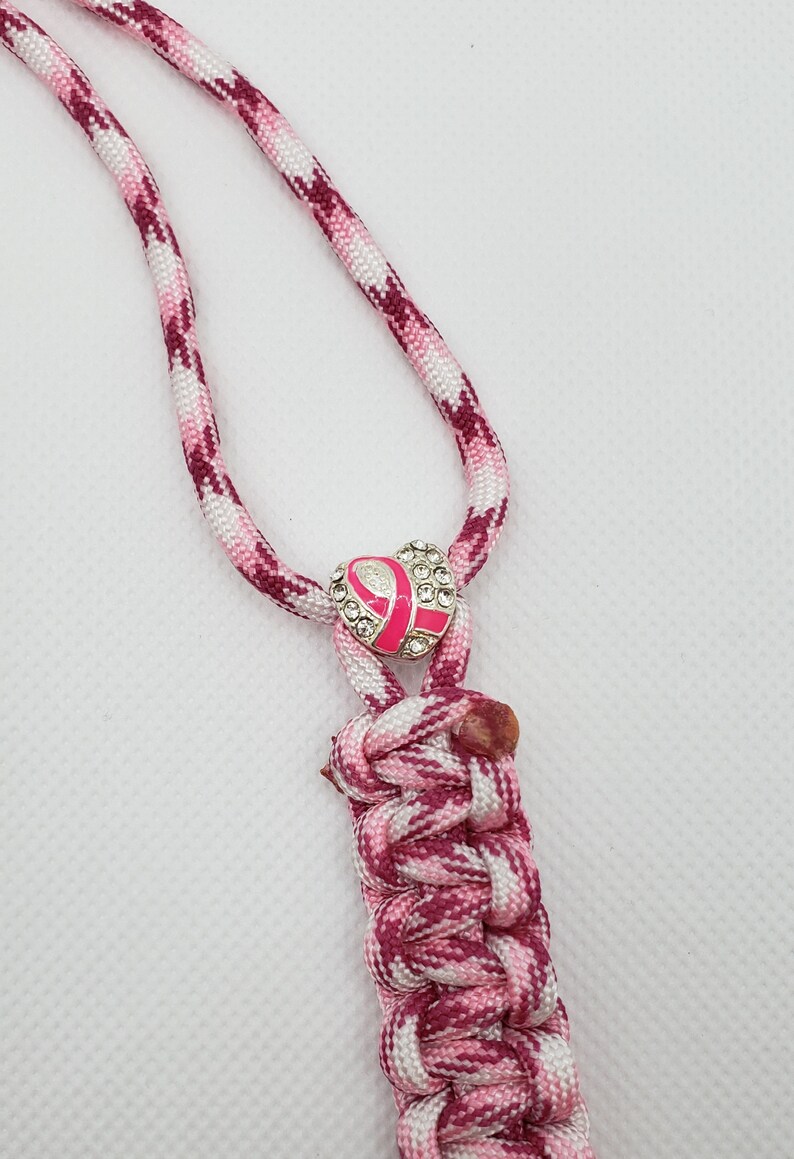May include: A pink and white braided lanyard with a pink ribbon charm. The charm is encrusted with rhinestones and has a pink ribbon in the center. The lanyard is made of a braided cord.