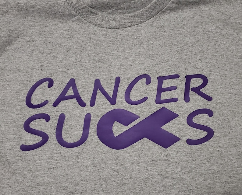 May include: A heather gray t-shirt with the phrase "CANCER SUCKS" in purple lettering and a purple awareness ribbon. The shirt has a crew neck and is made of a soft, comfortable material.