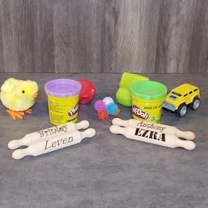 May include: A collection of children's toys, including two containers of Play-Doh, a yellow toy truck, a plush yellow chick, and two wooden rolling pins engraved with names. Also included are a red ball, a green toy, and colorful spiky balls.