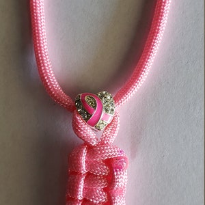 May include: Pink braided lanyard with a heart-shaped charm featuring a pink ribbon and sparkling accents. The lanyard is made of a woven, textured material. The charm is silver-toned.