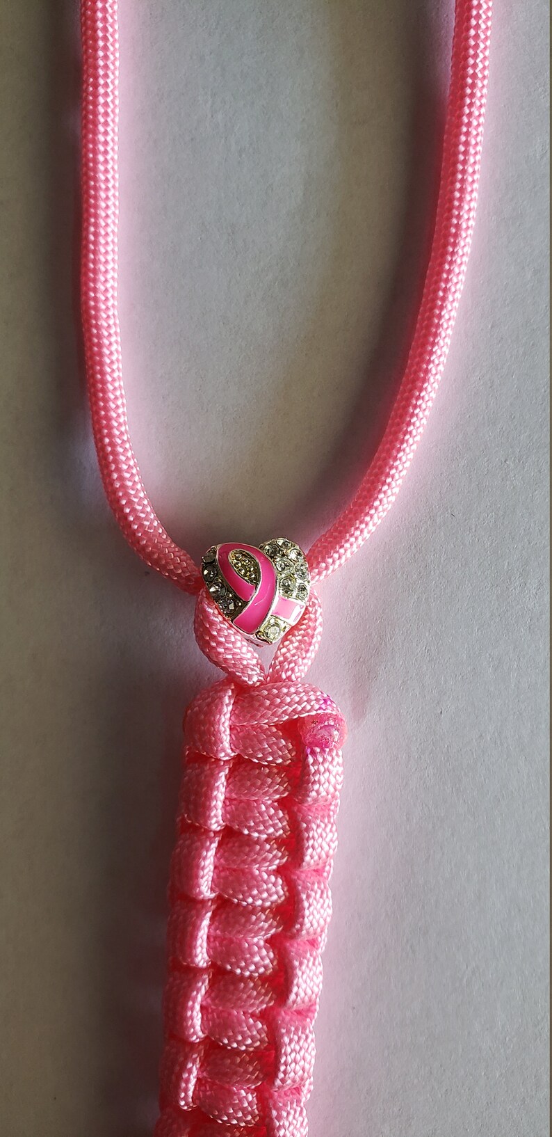 May include: Pink braided lanyard with a heart-shaped charm featuring a pink ribbon and sparkling accents. The lanyard is made of a woven, textured material. The charm is silver-toned.
